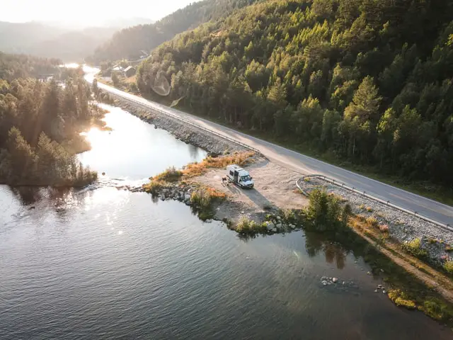 Vue aérienne d'un van blanc sur un parking au bord d'une route, d'un lac et d'une forêt de sapins.