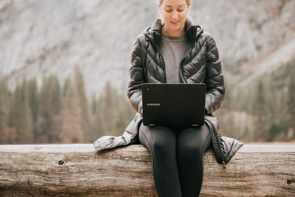 Femme en mentaux noire assise sur un tronc d'arbre travaillant sur son ordinateur portable.